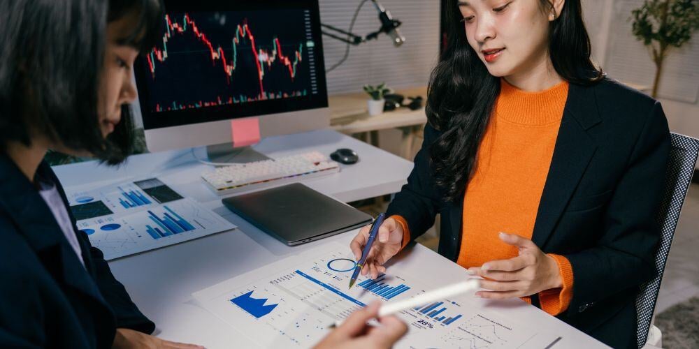 Two businesswomen collaborating on MSP ROI analysis with detailed graphs, charts, and a live market data screen in the background