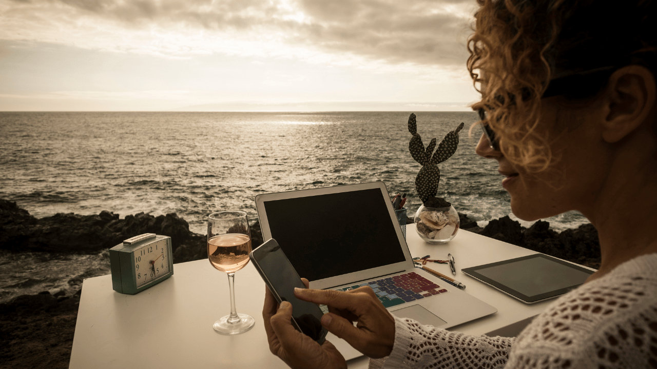 A person working on a laptop by the ocean, with a glass of wine, clock, and cactus on the table, enjoying a relaxed yet productive setting